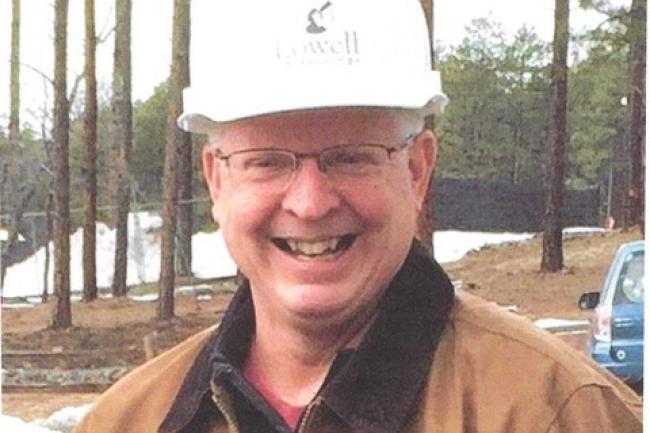 A photo of Kevin Schindler, a white man with a big smile, wearing a hard hat that reads "Lowell Observatory". He stands outside in front of pine trees and there is snow on the ground.