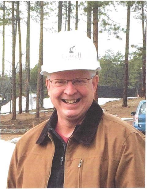 A photo of Kevin Schindler, a white man with a big smile, wearing a hard hat that reads "Lowell Observatory". He stands outside in front of pine trees and there is snow on the ground.